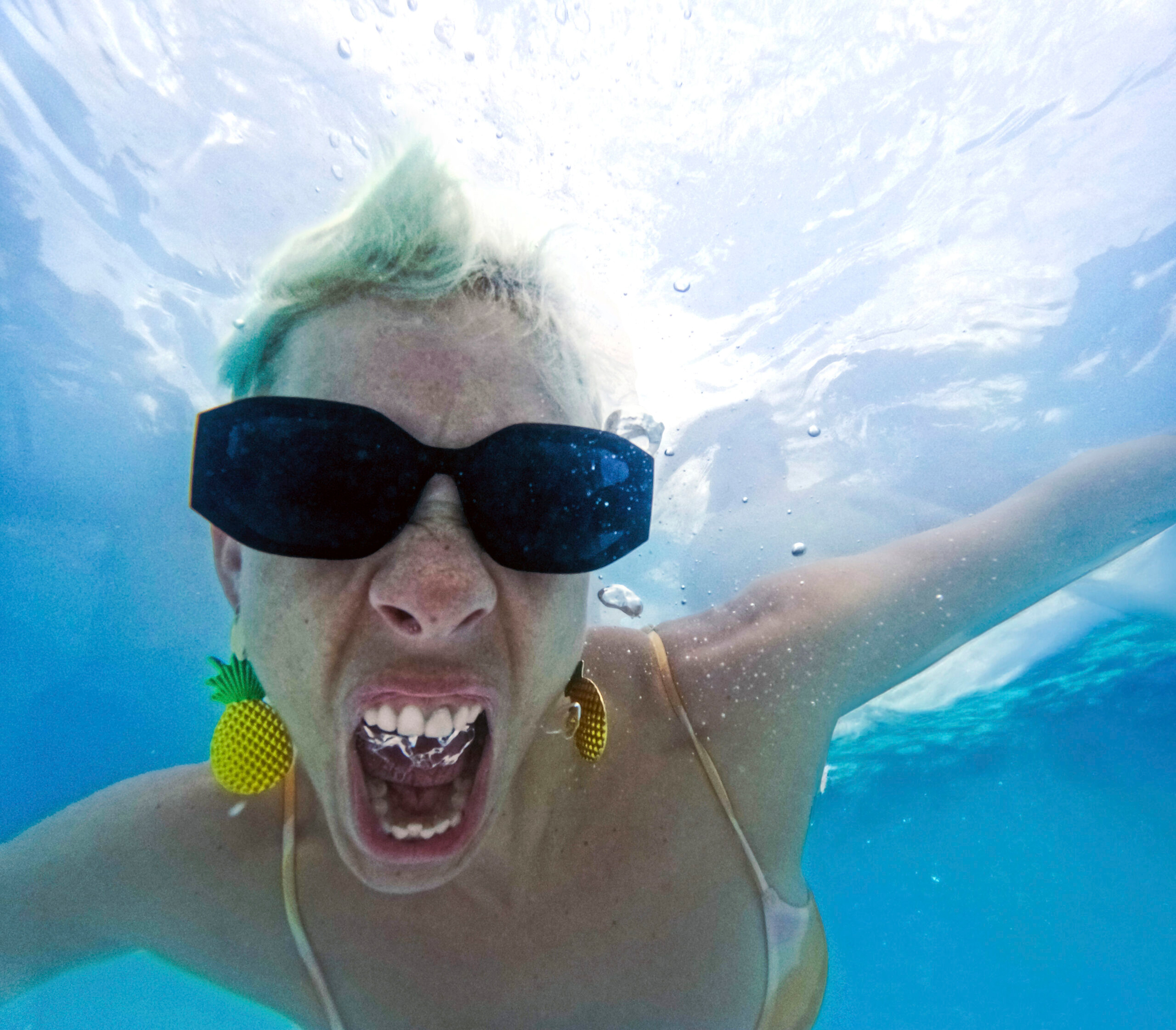 Krystal Lavenne underwater wearing black sunglasses and pineapple-shaped earrings, with short light hair, appears to be shouting or exhaling, creating bubbles around their open mouth. The water is bright blue with sunlight filtering from above, and one arm is extended out of frame.